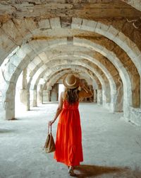 A traveller in a red dress and straw hat walking beneath a row of ancient stone arches