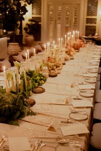 A long candlelit dinner table dressed with fruit, vegetables and greenery, with a place card and menu at every setting