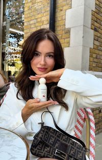 A creator outside a café, hand under the chin, holding up an Atelier Rebul bottle